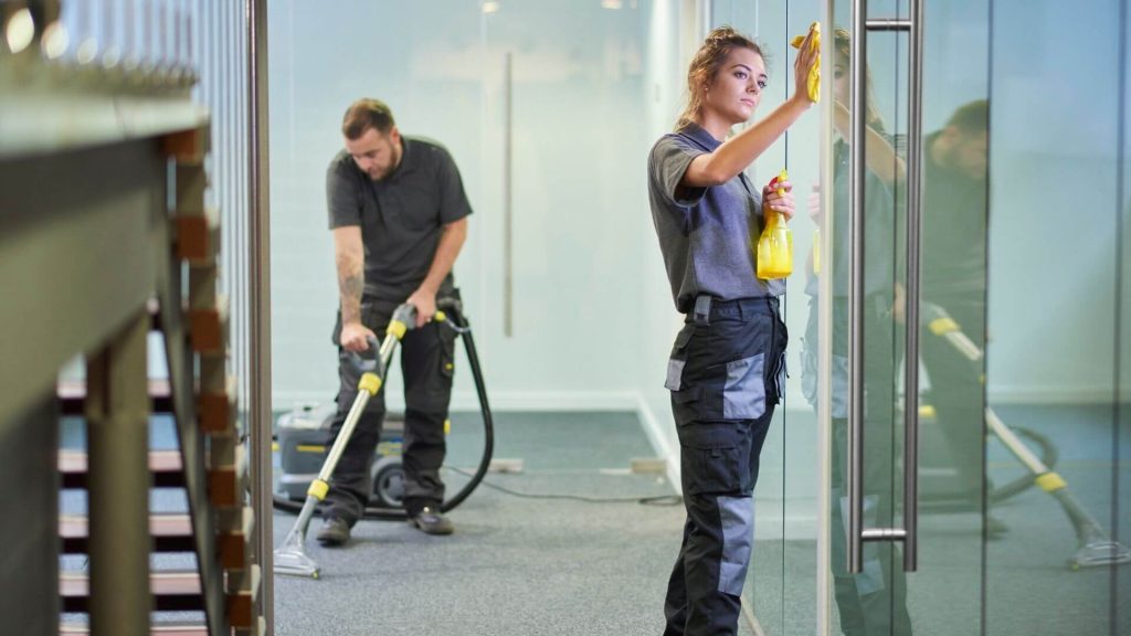 one person cleaning the glass wall and one person vacuuming the office carpet